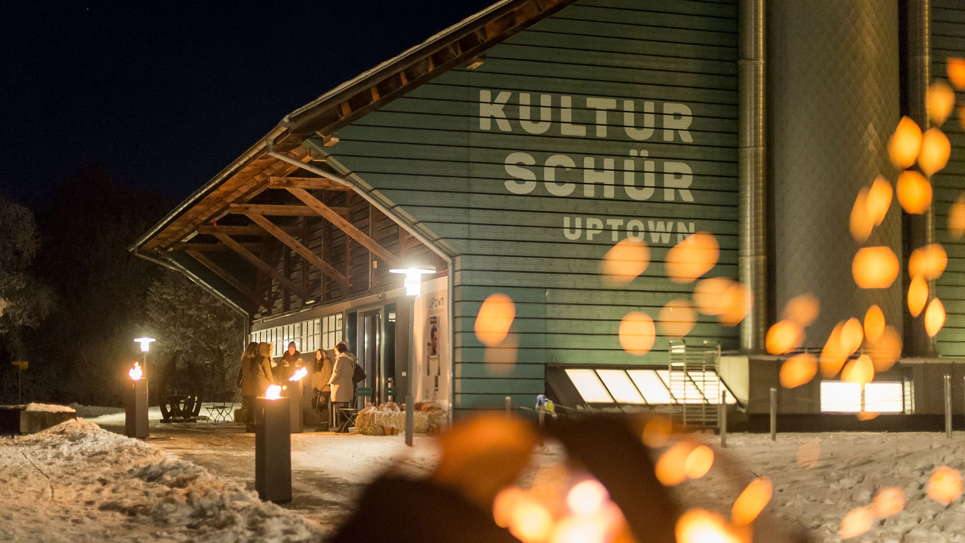 The Schüür event venue in winter, with a group of people in front of the entrance and cozy fire pits along the path.