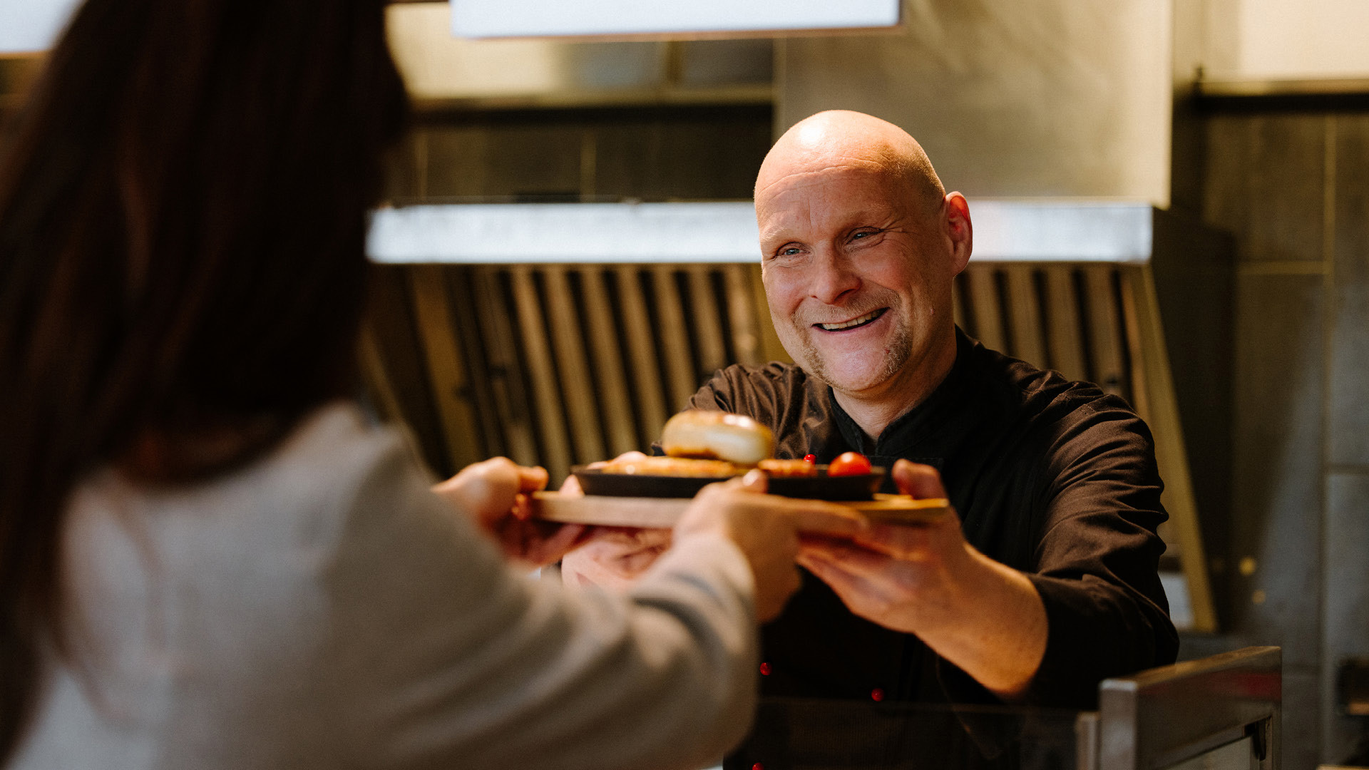 A cook in a black cooking jacket handing a plate to a guest from the buffet.